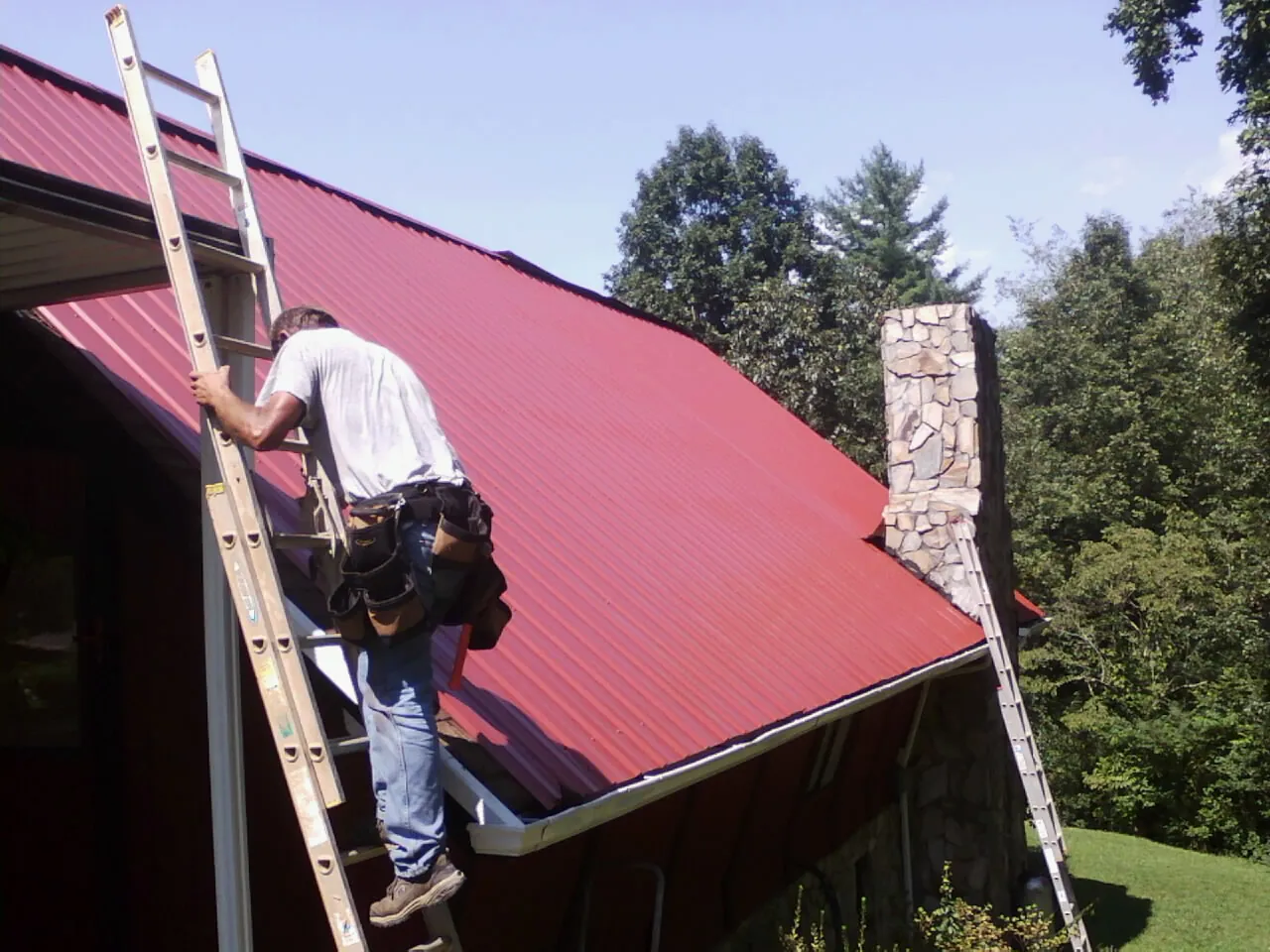 Roofer on a ladder beside a red metal roof and stone chimney — Soffit & Fascia Repair by Roofing Brothers in Canton